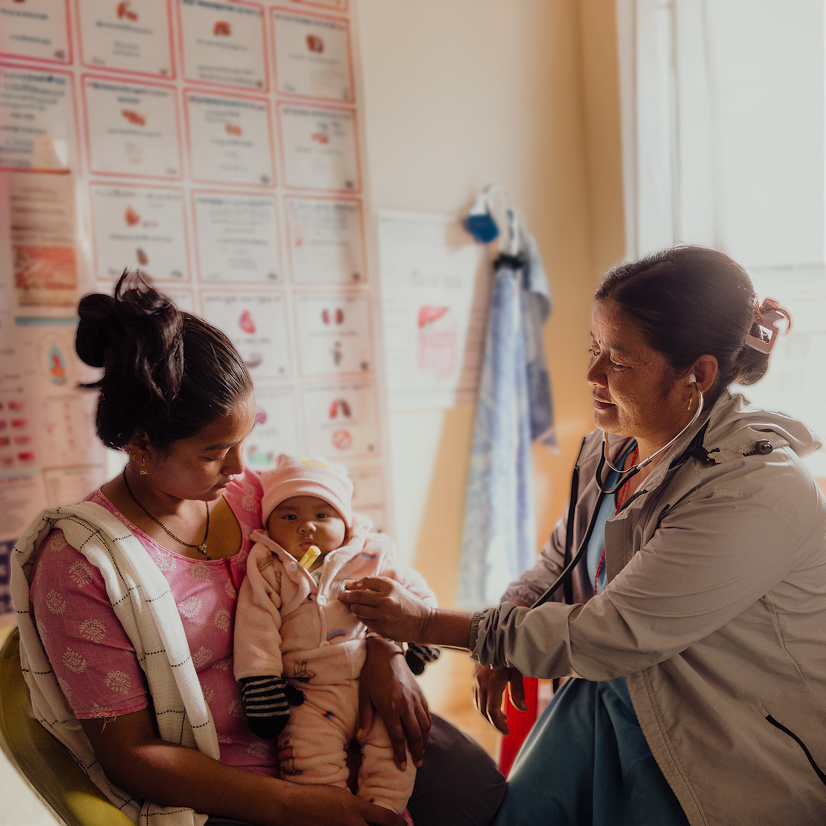A woman sits on a chair in a medical clinic, holding her baby girl as a female doctor listens to her heartbeat with a stethoscope.